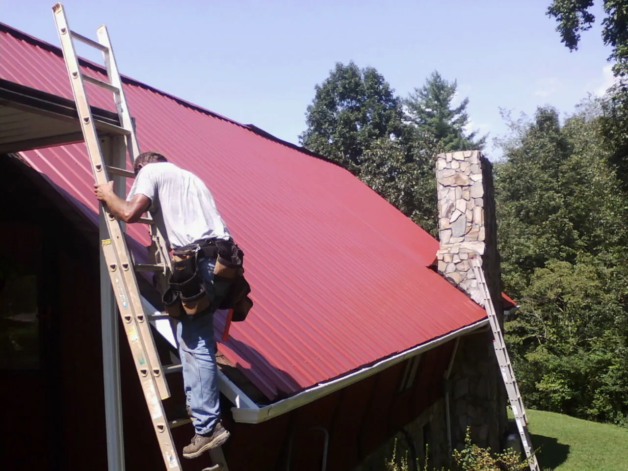 Roofer on a ladder beside a red metal roof and stone chimney — Soffit & Fascia Repair by Roofing Brothers in Manville